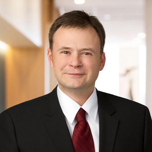 A man in a dark suit, white shirt, and red tie stands indoors in a brightly lit corporate law office hallway, facing the camera and smiling slightly.