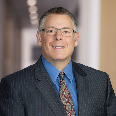 A man wearing glasses, a dark pinstripe suit, a blue shirt, and a patterned tie stands in the hallway of a corporate law office with blurred background lights.