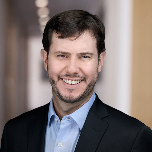 A man with short brown hair and a beard, wearing a blue shirt and dark blazer, smiles in a blurred indoor office hallway, representing one of the top law offices for chicago lawyers.