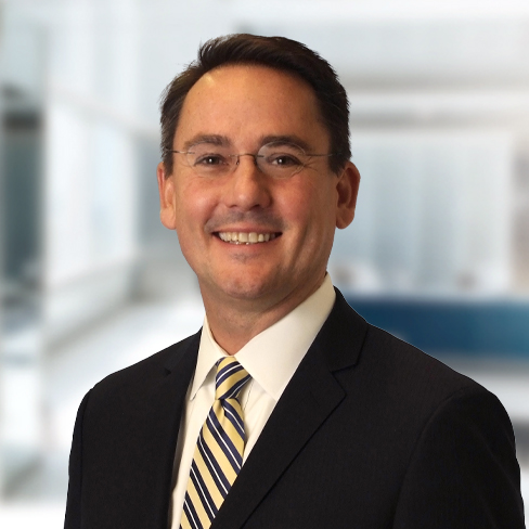 A man in a dark suit, white shirt, and striped tie smiles at the camera in a bright, blurred corporate law office, reflecting the professionalism of leading Chicago lawyers.