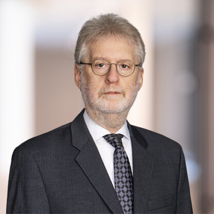 A middle-aged man with gray hair, glasses, and a beard wearing a dark suit, white shirt, and patterned tie stands against a blurred indoor background, evoking the professionalism of a corporate law office.