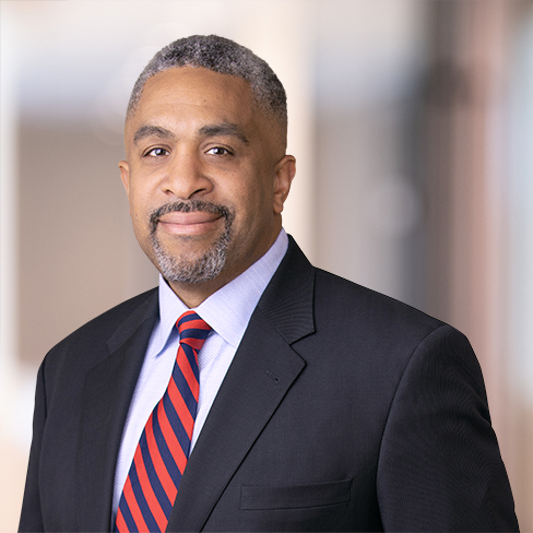 A man in a dark suit, light blue shirt, and red striped tie stands in a blurred law offices setting, facing the camera and smiling slightly.