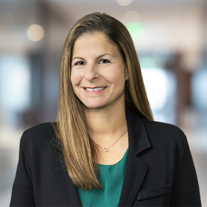 Woman with straight brown hair wearing a black blazer and green top, smiling, standing in a blurred indoor office setting, representing lawyers in Chicago who specialize in intellectual property law.