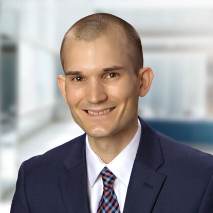 A man in a navy suit, white shirt, and plaid tie smiles at the camera against a blurred office background, representing chicago lawyers with expertise in intellectual property law.