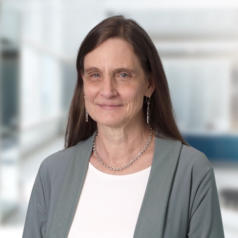 A middle-aged woman with long brown hair, wearing a gray cardigan, white top, silver necklace, and earrings, stands in a bright, blurred corporate law office.