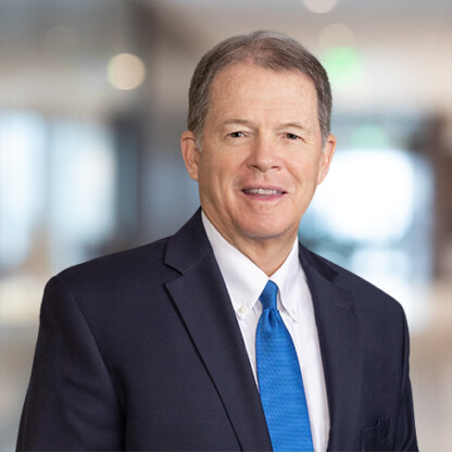 Man in a dark suit, white shirt, and blue tie standing indoors with a blurred corporate law office background.