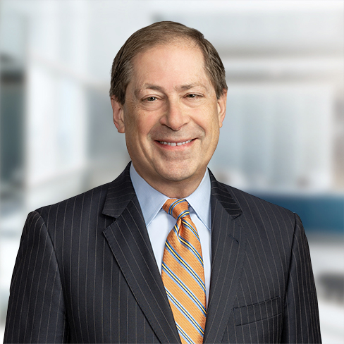 A middle-aged man in a dark pinstripe suit, light blue shirt, and striped tie smiles at the camera in a modern corporate law office setting.