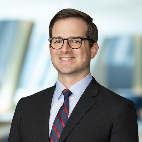 A man wearing glasses, a dark suit, a light blue shirt, and a striped tie stands in front of a blurred law offices background, smiling at the camera.