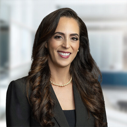 Woman with long brown hair wearing a black blazer and pearl necklace, smiling, posed in a modern, blurred law offices setting.