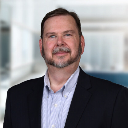 A middle-aged man with short brown hair, a beard, and mustache, wearing a dark suit jacket and light-striped shirt, stands in a modern office setting known for chicago lawyers specializing in intellectual property law.