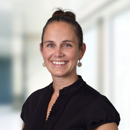 Woman with brown hair in a bun, wearing a black blouse and gold earrings, smiling at the camera in a brightly lit corporate law office setting.