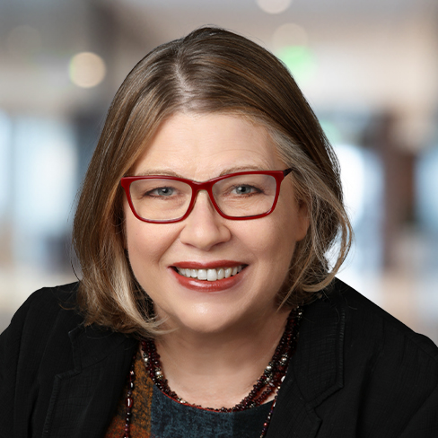 A woman with short, light brown hair and red glasses smiles at the camera. She is wearing a dark blazer and a multicolored top, with a blurred corporate law office background behind her.