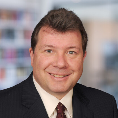 A man in a dark suit, white shirt, and patterned tie smiles at the camera with a blurred office background, embodying the professionalism of lawyers in Chicago specializing in intellectual property law.