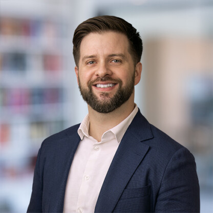 A man with short brown hair and a beard, wearing a navy blazer and light-colored shirt, smiles at the camera in a blurred law offices setting, reflecting the professionalism of lawyers in Chicago.