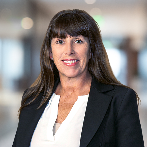 A woman with long brown hair, wearing a black blazer and white blouse, stands in a blurred corporate law office setting, looking at the camera and smiling.