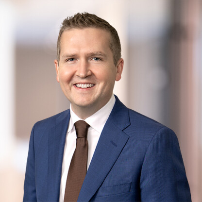 A man in a blue suit, white shirt, and brown tie smiles at the camera against a softly blurred corporate law office background.
