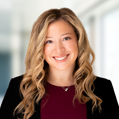 A woman with long, wavy blonde hair wearing a black blazer over a burgundy top, smiling at the camera in a brightly lit corporate law office, representing the professionalism of Chicago lawyers.