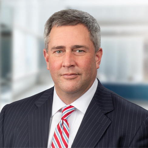 A middle-aged man with short gray hair wearing a pinstripe suit, white shirt, and red-striped tie, posing confidently against the blurred backdrop of a corporate law office in Chicago.