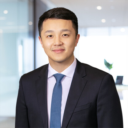 A man in a dark suit and blue tie stands in a modern, well-lit corporate law office, looking at the camera with a neutral expression.