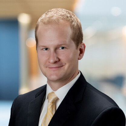 A man with short blond hair wearing a black suit, white shirt, and yellow striped tie poses for a professional headshot in a corporate law office against a blurred office background.