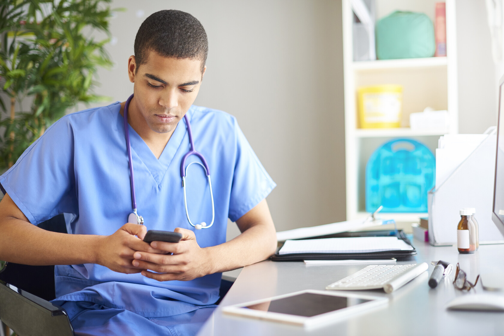 A healthcare professional in blue scrubs sits at a desk looking at a smartphone, with a stethoscope around his neck and medical supplies on the desk, reminiscent of the focus seen in busy Chicago lawyers or corporate law office settings.