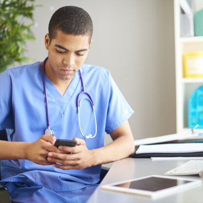 A healthcare professional in blue scrubs sits at a desk looking at a smartphone, with a stethoscope around his neck and medical supplies on the desk, reminiscent of the focus seen in busy Chicago lawyers or corporate law office settings.