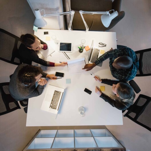 Four people sit around a white table with laptops, tablets, and papers, collaborating on work in a corporate law office setting.