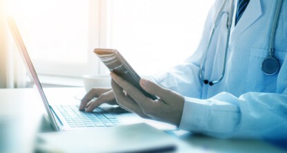 A doctor in a white coat using a smartphone and typing on a laptop at a desk with a stethoscope around their neck, seeking litigation support similar to chicago lawyers.