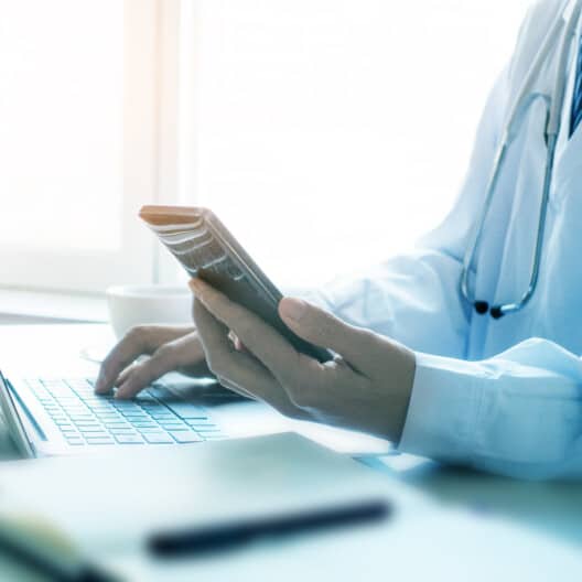 A doctor in a white coat using a smartphone and typing on a laptop at a desk with a stethoscope around their neck, seeking litigation support similar to chicago lawyers.