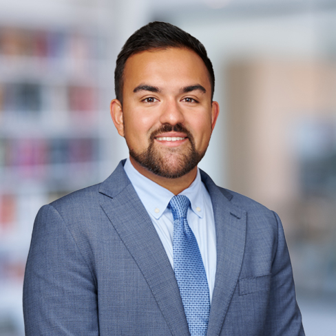 A man in a blue suit and tie stands in front of a blurred corporate law office background, facing the camera and smiling.