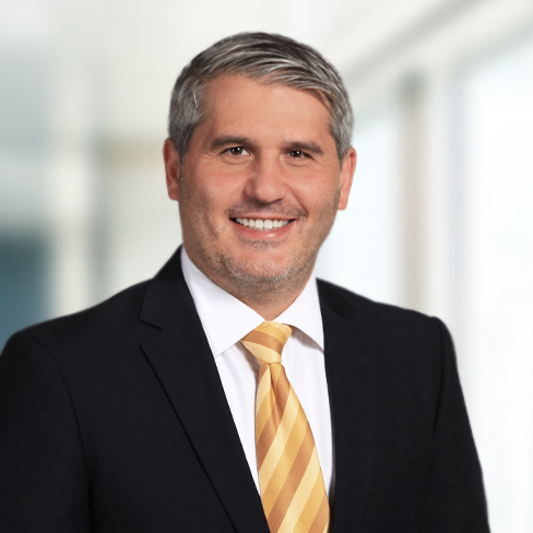 A man in a dark suit, white shirt, and yellow striped tie smiles at the camera in a bright, blurred office setting, embodying the professionalism of top Chicago lawyers.