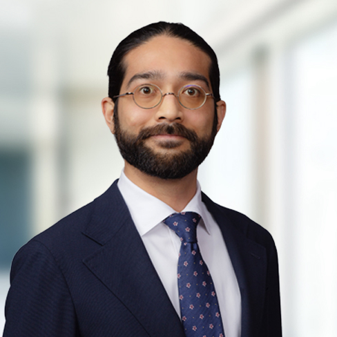 Man with dark hair and beard, wearing glasses, a blue suit, white shirt, and patterned tie, standing in a bright, blurred office setting—ideal for profiles of lawyers in Chicago or professionals in intellectual property law.