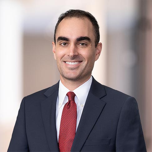 A man in a dark suit, white shirt, and red tie smiles at the camera against a blurred indoor background, reflecting the professional environment of a corporate law office.