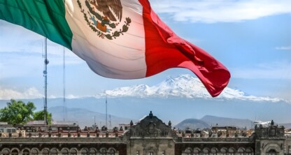 A large Mexican flag waves over historic buildings, with snow-capped mountains in the background and a prominent corporate law office under the clear blue sky.