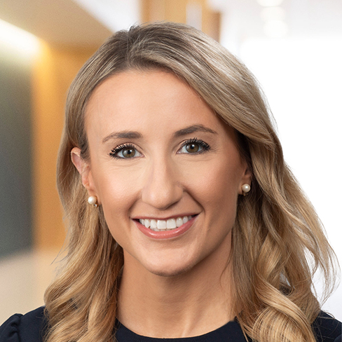 A woman with long blonde hair, wearing pearl earrings and a dark top, smiles at the camera in a brightly lit corporate law office.
