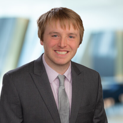 A young man with light brown hair, wearing a gray suit, light shirt, and tie, smiles at the camera with a blurred corporate law office in the background.