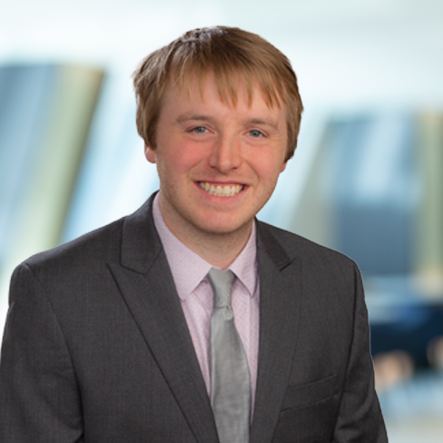 A young man with light brown hair, wearing a gray suit, light shirt, and tie, smiles at the camera with a blurred corporate law office in the background.