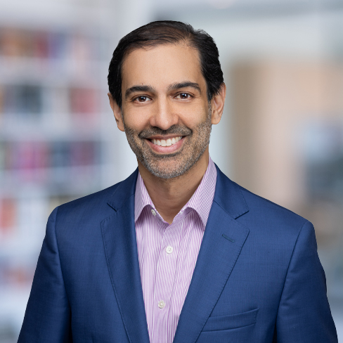 Man with short dark hair and beard wearing a blue suit jacket and light purple striped shirt, smiling in a softly blurred office setting, representing litigation support at one of the leading law offices.