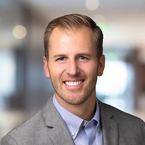 A man with short light brown hair and a trimmed beard, wearing a gray blazer and light blue shirt, smiles in a blurred indoor office setting typical of modern Chicago lawyers or corporate law offices.