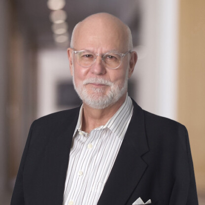 An older man with a white beard and glasses, wearing a striped shirt and dark blazer, stands in the hallway of a corporate law office with blurred background lights, embodying the professionalism of Chicago lawyers.