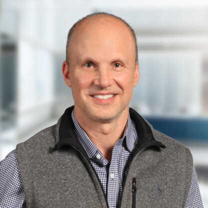 A middle-aged man with a shaved head, wearing a grey vest over a checkered shirt, stands in an office setting—ideal for professionals like chicago lawyers or those providing litigation support.