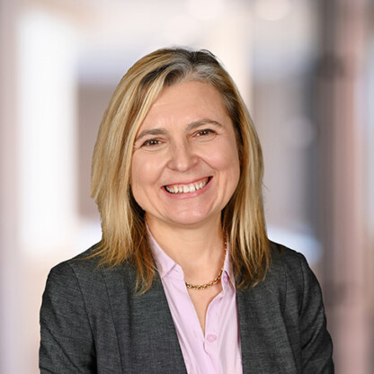 A woman with blonde hair wearing a gray blazer and light pink blouse smiles in front of a blurred indoor background, reflecting the professional atmosphere of corporate law offices.