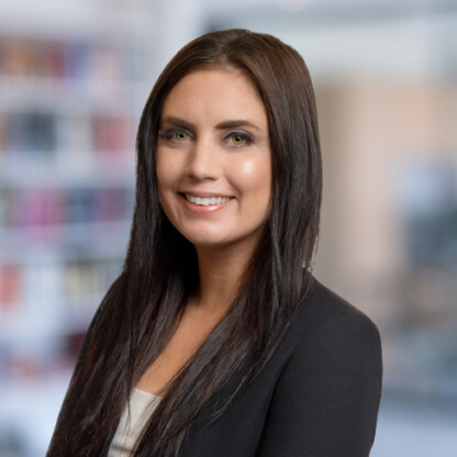 A woman with long dark hair and a black blazer smiles while standing in front of a blurred office, reflecting the professional atmosphere of a corporate law office.