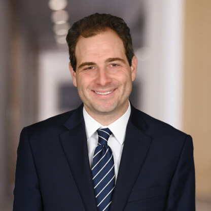 A man in a dark suit and striped tie stands smiling in a blurred hallway, reflecting the professional atmosphere of law offices specializing in intellectual property law.