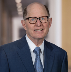 A middle-aged man in a blue suit, white shirt, and blue tie, wearing glasses, smiles at the camera in an indoor setting with blurred background—reflecting confidence often seen in law offices specializing in intellectual property law.