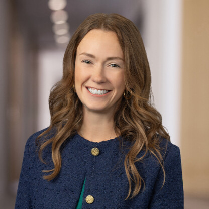 A woman with wavy brown hair, wearing a blue jacket and green top, smiles at the camera in a brightly lit law office hallway, offering a welcoming presence for clients seeking litigation support.