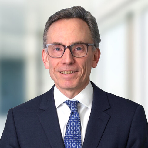 A man in a dark suit, white shirt, and patterned blue tie smiles at the camera, wearing glasses; the bright, blurred indoor setting hints at a bustling corporate law office.