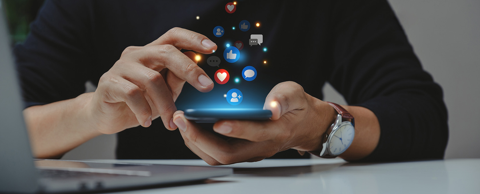 Person using a smartphone with social media icons like likes, hearts, and comments appearing above the screen. A laptop is visible on the table—perfect for chicago lawyers staying connected or working on intellectual property law cases.