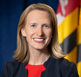 Woman with shoulder-length blonde hair wearing a dark blazer and red top, smiling in front of a Maryland state flag, offers trusted litigation support and expertise akin to top lawyers in Chicago.