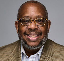 A man with glasses, a short beard, and a mustache, wearing a brown checked blazer and striped shirt, smiles at the camera against a plain gray background—exuding the confidence often seen in top Chicago lawyers.